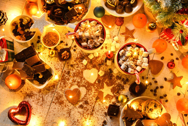 sweet food top view background for merry christmas or new year holiday decoration with night illumination - chocolate candies, tangerines, cookies, marshmallow and cocoa latte on white wood - Foto, afbeelding