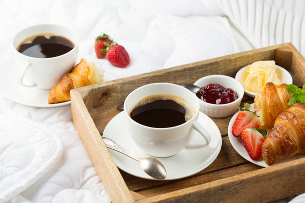 Desayuno en la cama, bandeja con tazas de café, cruasanes frescos, mermelada, fresas. Concepto de luna de miel. Temprano en el hotel. Enfoque selectivo, espacio de copia. - Foto, imagen