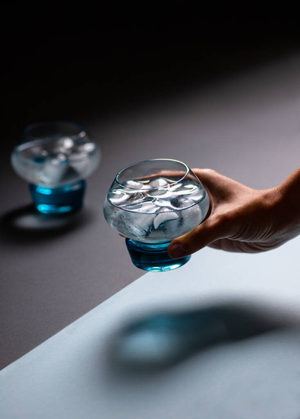 女性の手は黒と青の背景に水と氷の青いガラスを保持します。冷たいさわやかな飲み物。最小限の構成 - 写真・画像