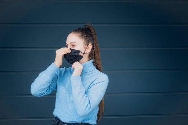 mladá dívka nasadí černou ochrannou masku na šedé pozadí, žena v modrém po kolena, místo pro reklamní nápis, osobní ochranné prostředky proti koronaviru - Fotografie, Obrázek