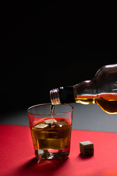 Whisky y piedras de whisky en un vaso sobre fondo rojo y negro. Una bebida alcohólica se vierte de una botella en un vaso. Composición minimalista - Foto, imagen