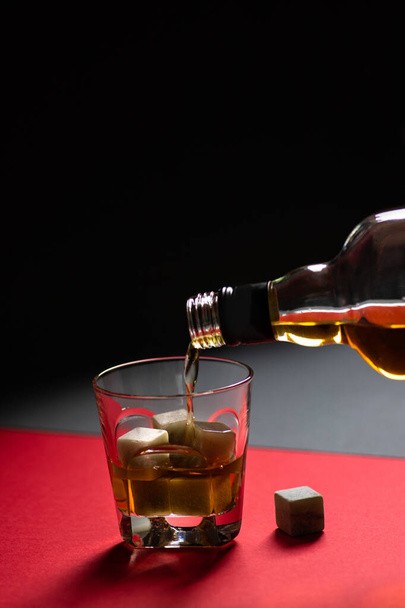 Whiskey und Whiskeysteine in einem Glas vor rotem und schwarzem Hintergrund. Ein alkoholisches Getränk wird aus einer Flasche in ein Glas gegossen. Minimalistische Komposition - Foto, Bild