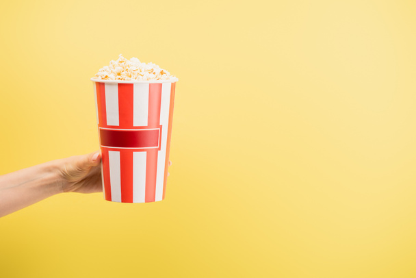 partial view of woman holding bucket of delicious popcorn isolated on yellow, cinema concept - Fotó, kép