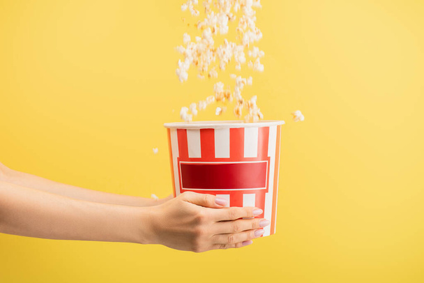 tasty popcorn falling into striped bucket in female hands isolated on yellow, cinema concept - Photo, Image