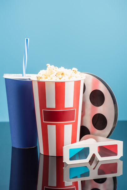 bucket with popcorn, cup with soda, film bobbin and 3d cinema glasses on glossy surface isolated on blue - Photo, Image