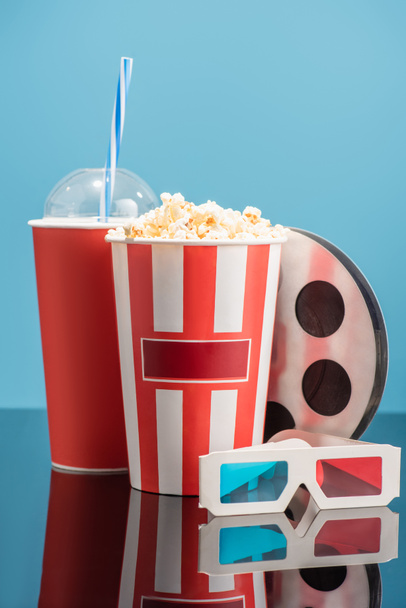 cup of soda, bucket with popcorn, 3d glasses and film bobbin on glossy surface isolated on blue, cinema concept - Photo, Image