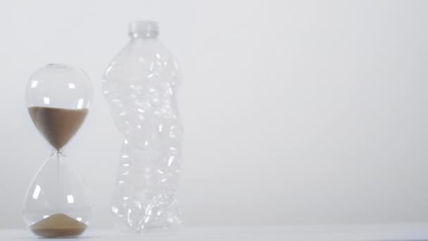Una amplia toma de estudio de una botella de plástico de un solo uso aplastada junto a un temporizador de arena sobre un fondo blanco - Metraje, vídeo