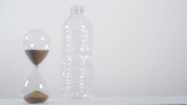 Egy széles stúdiófelvétel egy egyszer használatos műanyag palackról egy homokóra mellett, fehér alapon - Felvétel, videó