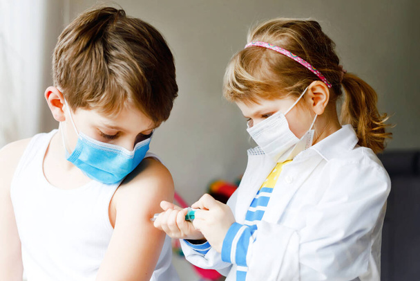 Little girl makes injection to brother, school kid boy. Children, siblings with medical mask playing doctor, holding syringe with vaccine. Coronavirus covid vaccination concept. Kids play role game - Photo, Image