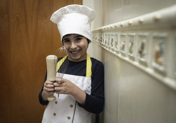 Usmívající se chlapec pózuje v kuchyni svého domu s kuchařskou uniformou Konceptuální životní styl, domácí život, porod a nové normální - Fotografie, Obrázek