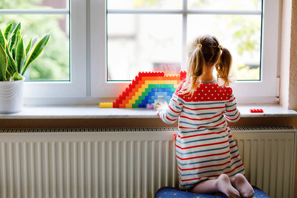 Χαριτωμένο μικρό κοριτσάκι από το παράθυρο δημιουργούν ουράνιο τόξο με πολύχρωμα πλαστικά μπλοκ κατά τη διάρκεια πανδημίας καραντίνα coronavirus. Παιδιά φτιαγμένα και ζωγραφισμένα ουράνια τόξα σε όλο τον κόσμο ως σημάδι. - Φωτογραφία, εικόνα