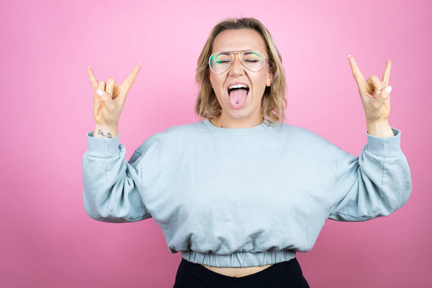 Молодая белая женщина в толстовке на розовом фоне кричит с сумасшедшим выражением, делая рок-символ с поднятыми руками - Фото, изображение
