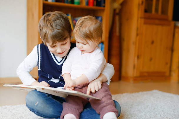 School kid boy reading book for little toddler baby girl, Two siblings sitting together and read books. Beautiful lovely family in love, cute baby and child having fun at home, indoors. - Foto, immagini