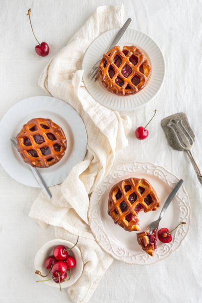 Vysokoúhlý pohled na tři třešňové koláče s vidličkami a čerstvými třešněmi na bílých talířích, bílým pozadím a stříbrným nádobím - Fotografie, Obrázek
