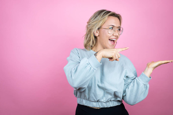 Jonge blanke vrouw draagt sweatshirt over roze achtergrond verrast, tonen en wijzen iets dat op haar hand - Foto, afbeelding