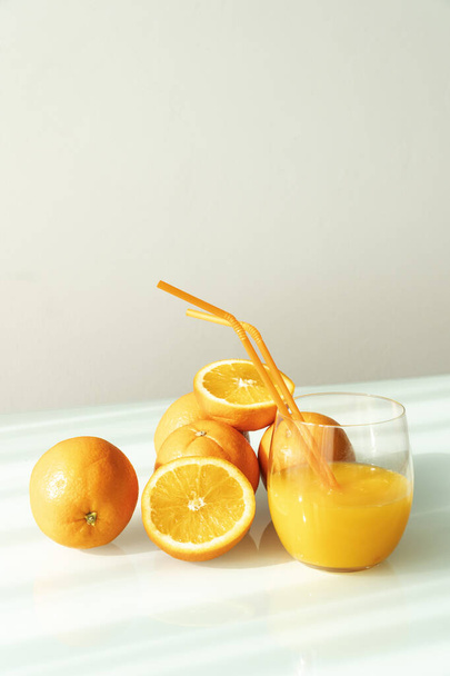 Jugo de naranja en un vaso rodeado de naranjas naturales con un fondo blanco claro al sol natural - Foto, imagen