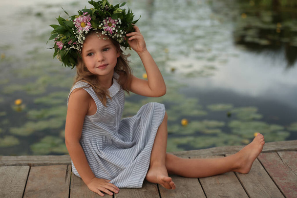 Cute little girl wearing wreath made of beautiful flowers on pier near pond - Foto, Imagen