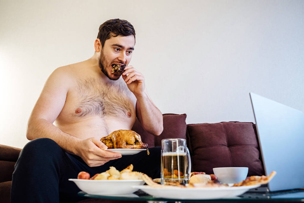 portrait of a caucasian fat man eating chicken grill on the sofa. looking at camera, at home. Unhealthy lifestyle concept - Фото, зображення