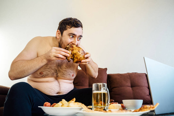 portrait of a caucasian fat man eating chicken grill on the sofa. looking at camera, at home. Unhealthy lifestyle concept - Fotoğraf, Görsel