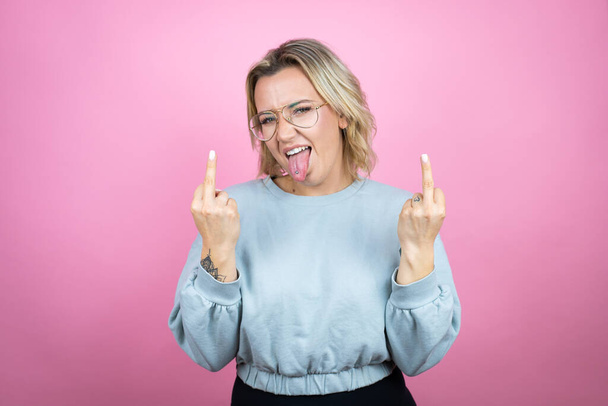 молодая белая женщина в толстовке на розовом фоне показывает средний палец делает трахнуть вас плохое выражение лица, провокация и грубое отношение. Крики возбужденных - Фото, изображение