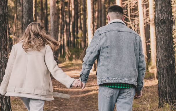 Ένα κορίτσι και ένας άντρας κρατιούνται χέρι-χέρι περπατώντας και διασκεδάζοντας σε ένα πευκοδάσος. - Φωτογραφία, εικόνα