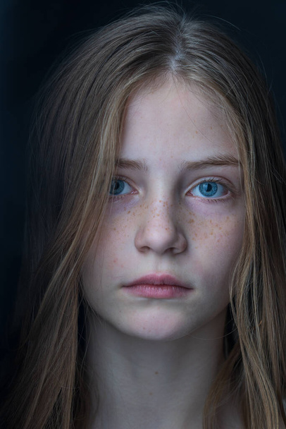 Charming blond young girl with freckles and blue eyes on a black background, indoors, close up portrait - Photo, Image