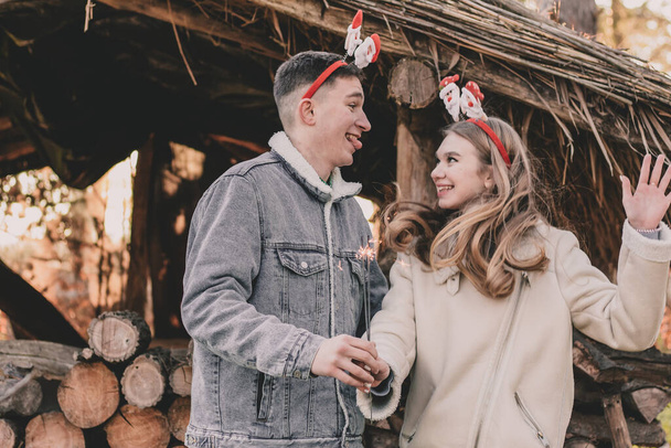 Ένα ζευγάρι για την εμφάνισή τους είναι ντυμένοι με στεφάνια Πρωτοχρονιάς στα κεφάλια τους, κρατώντας μια σπίθα στα χέρια τους και χαμογελώντας ο ένας στον άλλο στο φόντο ενός κιόσκι φτιαγμένο από καυσόξυλα - Φωτογραφία, εικόνα
