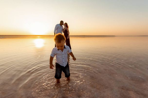 Portrait of little mixed race boy. Kissing parents on background. Happy multi ethnic family at the beach. Beautiful sunset - Photo, Image