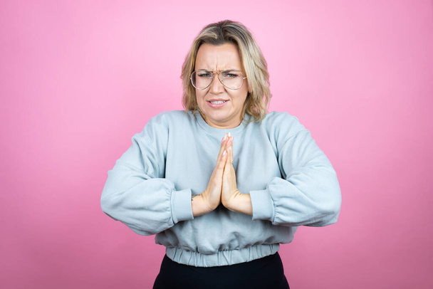 Jonge blanke vrouw draagt sweatshirt over roze achtergrond bedelen en bidden met de handen samen met hoop uitdrukking op het gezicht zeer emotioneel en bezorgd - Foto, afbeelding