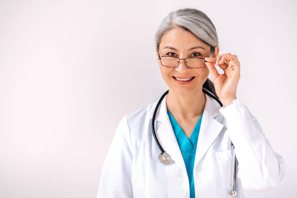 Retrato de cerca de la atractiva doctora caucásica en uniforme médico y estetoscopio sobre un fondo blanco, mira a la cámara y sonríe. Concepto sanitario - Foto, Imagen