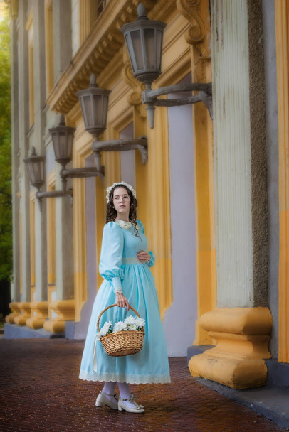 girl in a blue vintage dress with a basket of flowers near a beautiful mansion. - Foto, afbeelding