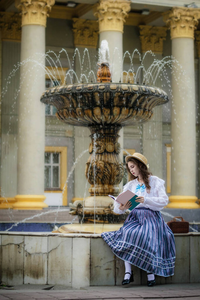Girl in a white dress reads a book while sitting at fountain against the background of an old building. - Foto, afbeelding