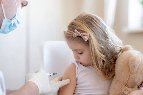 Immunization for children concept. Happy little cute blonde girl holding a toy and getting a flu shot not afraid of the syringe needle. Doctor injecting brave child with Covid-19 vaccine at clinic or - Photo, image