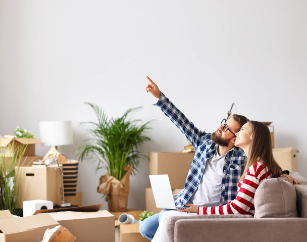 Side view of optimistic married young couple: man with laptop pointing up and discussing design of new apartment with wife while sitting together on sofa and browsing internet catalog of furniture in room with packed boxes - Photo, Image