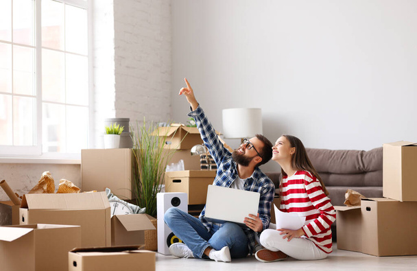 Full body of happy married couple: young man with laptop on knees pointing up while sitting with wife among packed stuff and discussing ideas about interior design of new home - Photo, Image