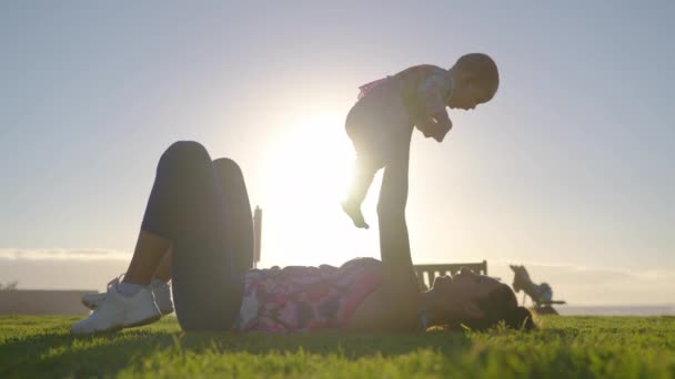 Nuori äiti pitää sylissään ja leikkii tyttövauvan kanssa meren rannalla auringonlaskun aikaan loman aikana.. - Materiaali, video