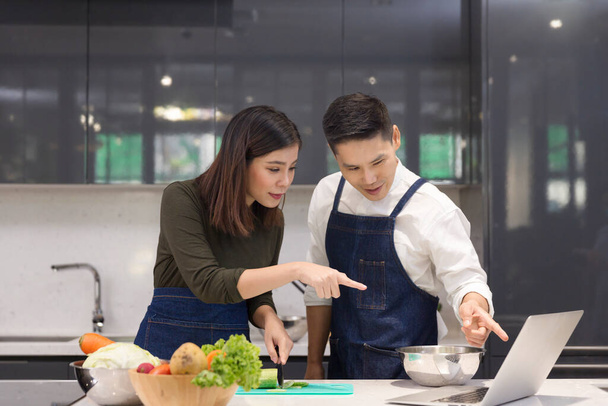 家庭のキッチンで一緒にアジアのカップル料理。女性は野菜をスライスし、ラップトップコンピュータで調理します。若いカップルはキッチンで野菜を切るのを手伝っています. - 写真・画像