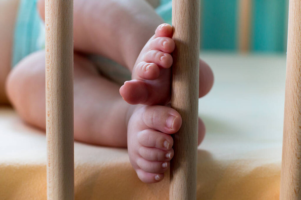 Τα πόδια του μωρού κοντά, λεπτομέρεια από τα πόδια του βρέφους με μικρά δάχτυλα και καρφιά στα μαλακά μικρά πόδια, το βρέφος στο κρεβάτι ξαπλωμένο και παίζοντας με τα πόδια στις κολώνες της κούνιας - Φωτογραφία, εικόνα