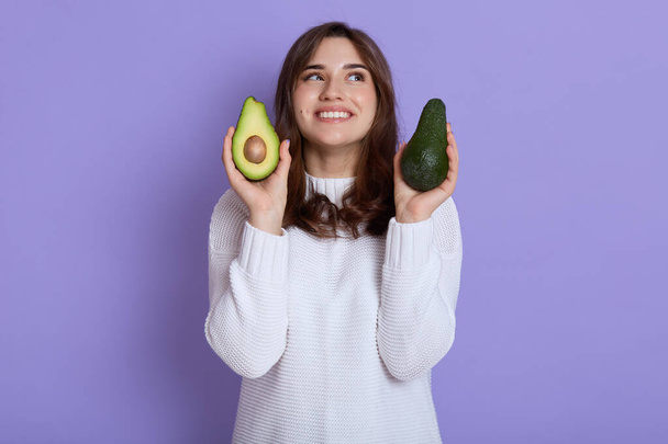 Happy woman standing isolated over lilac background and holding two halves of avocado, looking smiling up, wearing casual white sweater, expressing positive emotions. - Foto, Bild