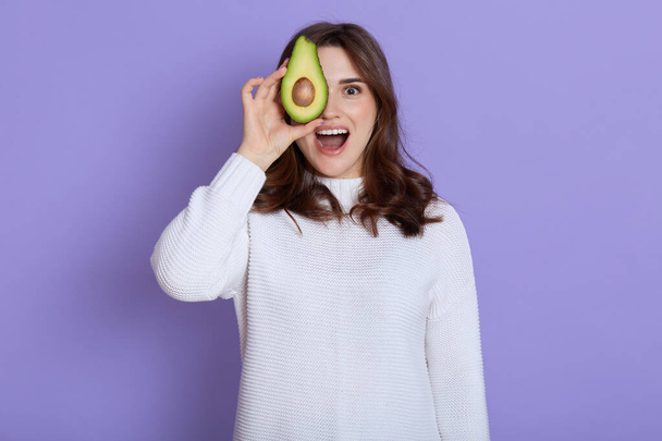 Happy excited young woman holding avocado half, covering eye with healthy fruit and keeping mouth opened, yelling something while standing against lilac wall, dresses white jumper. - Photo, image