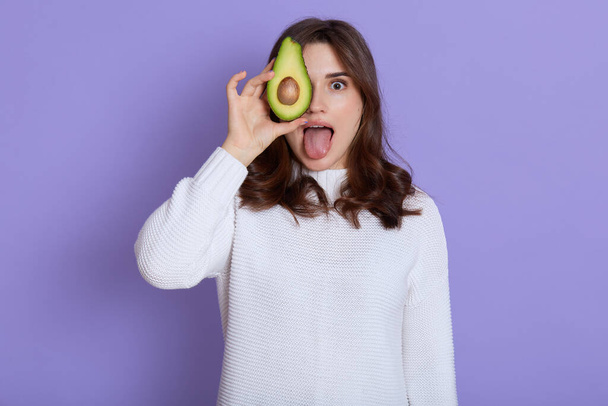 Young adorable european female in white casual clothing covering her eye with half of avocado, keeps healthy eating, looks at camera and showing tongue, poses isolated over lilac background. - Photo, image