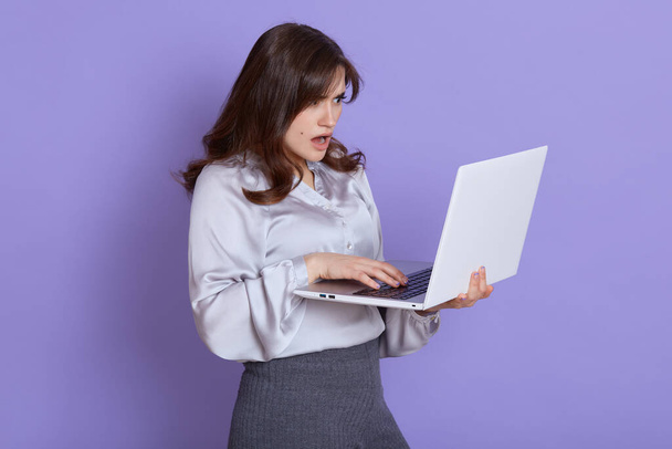 Young woman having trouble with laptop, posing against lilac wall and holding notebook with shocked and astonished facial expression, has problem with her online work, wearing elegant clothing. - Фото, зображення