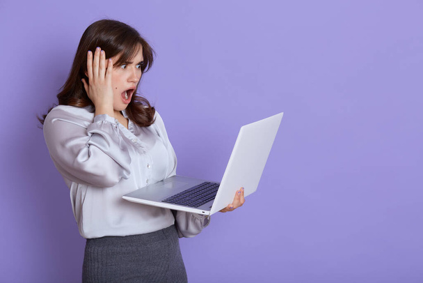 Attractive nervous young business woman with laptop in hands, looking at screen of device with shocked expression, touching her head with hand, keeps mouth widely opened, stands against lilac wall. - Foto, Imagen