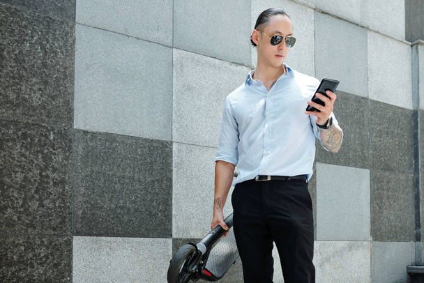 Beau jeune entrepreneur en lunettes de soleil debout à l'extérieur avec scooter électrique et lecture de SMS sur smartphone - Photo, image