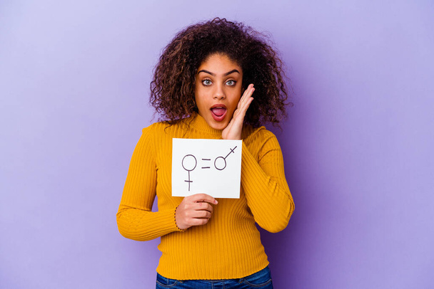 Junge Afroamerikanerin mit einem Plakat zur Gleichberechtigung der Geschlechter auf violettem Hintergrund überrascht und schockiert. - Foto, Bild
