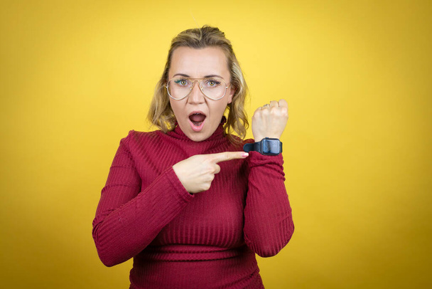 Joven mujer caucásica vistiendo casual camiseta roja sobre fondo amarillo sorprendido y señalando su reloj porque es tarde - Foto, Imagen