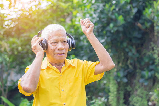 Старший мужчина стоит в беспроводных наушниках, слушая любимую песню, и улыбается, стоя в саду. Пространство для текста. Концепция пожилых людей и отдыха. - Фото, изображение