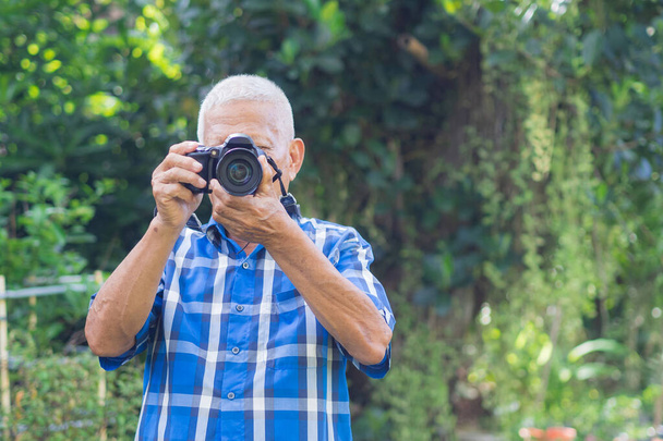 Hombre mayor filmando una foto con una cámara digital en el jardín. Un anciano lleva una camisa azul, feliz cuando usa una cámara. Concepto de personas mayores y fotografía. - Foto, imagen