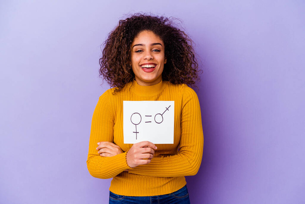 Giovane donna afroamericana in possesso di un cartello di uguaglianza di genere isolato su sfondo viola ridere e divertirsi. - Foto, immagini