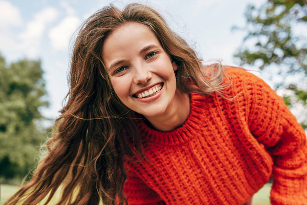 Candid portrait of a cheerful young woman wearing an orange sweater spending time in the park. The pretty female has a joyful expression, resting outdoors on nature background. - Foto, Imagen